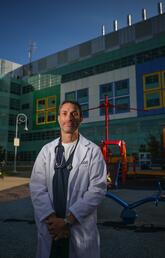 A man in a white coat stands in front of a hospital