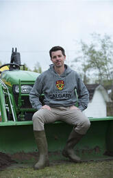 A man sits on a tractor while wearing a UCalgary sweatshirt