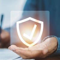 The hand of a man holding a icon with a shield with a checkmark inside it.