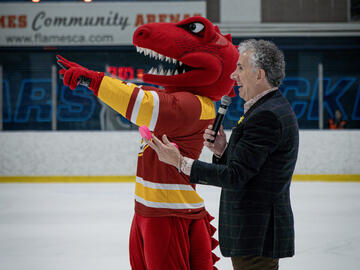 A red dinosaur mascot stands next to a man speaking into a microphone and speaking to a crowd