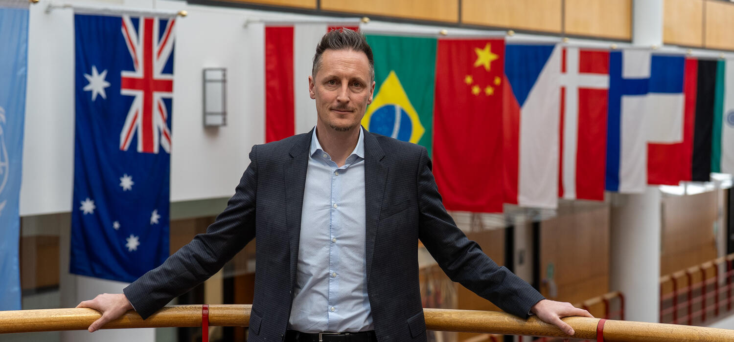 A man in a suit stands in front of multiple international flags