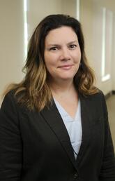 A woman with long dark hair and a suit looks at the camera