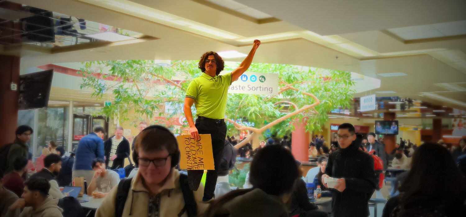 A man stands on a table in a crowded cafeteria while holding his fist in the air